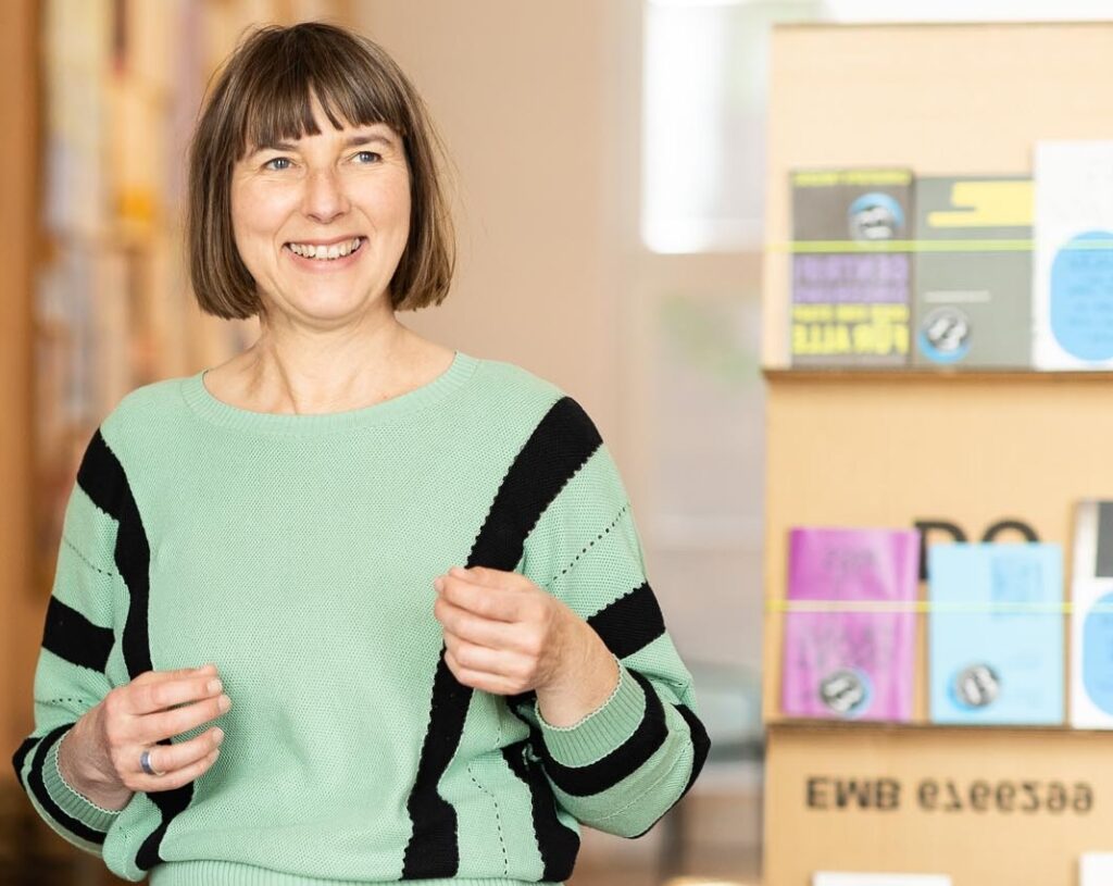 Ich stehe in einem Ausstellungsraum und erkläre etwas. Ich trage einen hellgrünen Pullover mit schwarzen Längsstreifen an den Seiten. Rechts von mir ist ein Ständer mit Flyern.